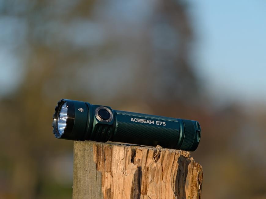 A teal Acebeam E75 flashlight sittting on top of a post with a blurry background