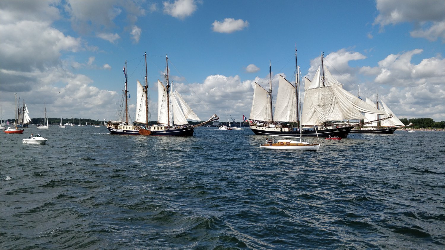 Photo of two three-mast sailing ships with full sails on a sunny day, surrounded by several smaller boats.